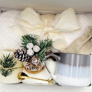 May include: A white gift box filled with a white fluffy blanket, a white mug with a grey rim, a small potted evergreen plant with pine cones, a small golden spoon, and a hessian bag.