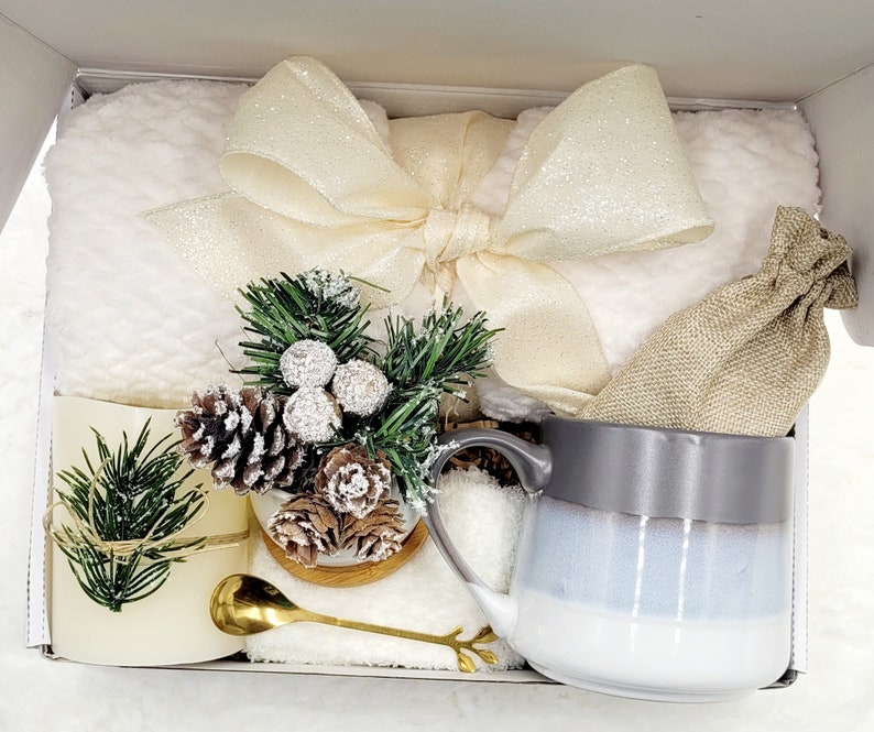 May include: A white gift box filled with a white fluffy blanket, a white mug with a grey rim, a small potted evergreen plant with pine cones, a small golden spoon, and a hessian bag.