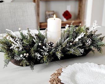 May include: A dark brown wooden dough bowl centerpiece filled with artificial frosted evergreen branches, pinecones, and white berries. A single white pillar candle sits in a clear glass holder in the center of the arrangement, emitting a warm glow.