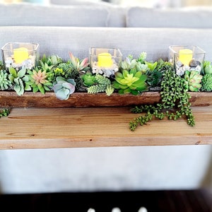 以下が含まれることがあります： 木製のトロフに多肉植物と、白い石と灯りがともったろうそくが入ったガラスのキャンドルホルダーが3つ置かれています。トロフは木製のテーブルの上に置かれています。