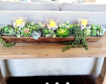 May include: A wooden trough filled with succulents and three glass candle holders with white stones and lit candles. The trough is sitting on a wooden table.