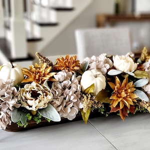 May include: A long, narrow wooden dough bowl centerpiece filled with artificial flowers and pumpkins. The arrangement features white pumpkins, light pink hydrangea clusters, golden-brown dahlias, green foliage, and small sprigs of dried-look filler.