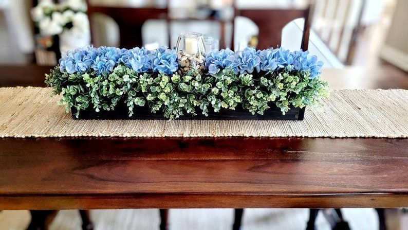 May include: A wooden box centerpiece with blue hydrangeas and green foliage. The centerpiece is on a woven table runner.