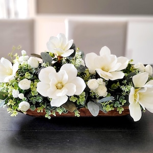 May include: A wooden bowl filled with a floral arrangement of white magnolia flowers and greenery. The bowl is sitting on a dark wood table.