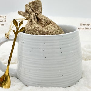May include: A white clay mug with a gold spoon and a burlap bag inside. The mug has a handle and a textured, ridged design. The gold spoon has a leaf-like design at the top. The burlap bag is tied with a string.