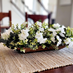 May include: A wooden dough bowl filled with a white flower arrangement. The flowers are surrounded by green leaves and are arranged in a long, rectangular shape. The bowl is sitting on a brown table with a woven placemat.