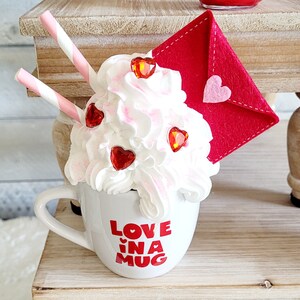 May include: White ceramic mug with a red and white heart design and the text "Love in a Mug". The mug is topped with whipped cream, red heart sprinkles, and a red felt envelope.