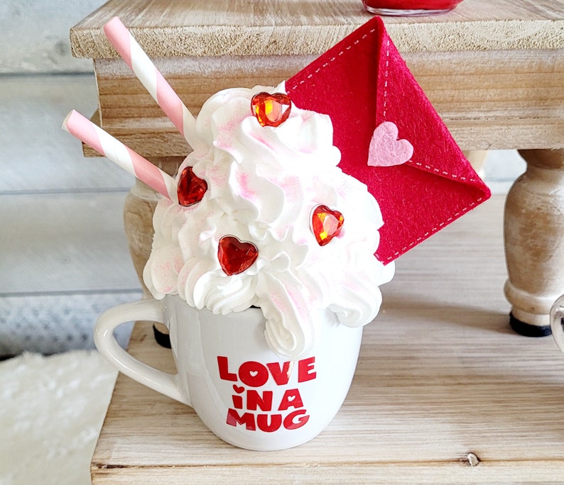 May include: White ceramic mug with a red and white heart design and the text "Love in a Mug". The mug is topped with whipped cream, red heart sprinkles, and a red felt envelope.