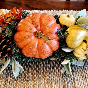 May include: A fall-themed centerpiece with a large orange pumpkin, gourds, pinecones, sunflowers, and white flowers. The centerpiece is arranged on a brown woven table runner.