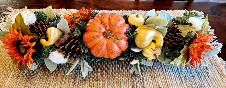 May include: A fall-themed centerpiece with a large orange pumpkin, gourds, pinecones, sunflowers, and white flowers. The centerpiece is arranged on a brown woven table runner.