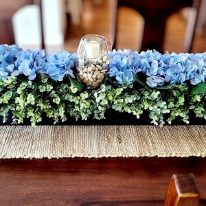 May include: A blue and green floral centerpiece in a black planter. The centerpiece features blue hydrangeas and green foliage. A glass vase with white stones is in the center.