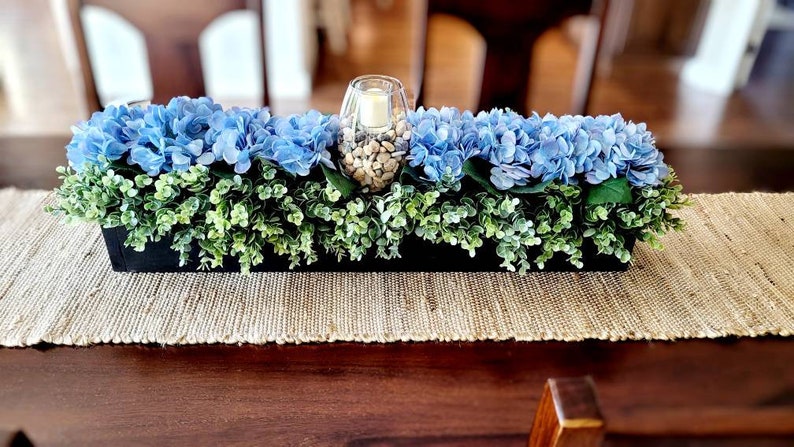 May include: A blue and green floral centerpiece in a black planter. The centerpiece features blue hydrangeas and green foliage. A glass vase with white stones is in the center.