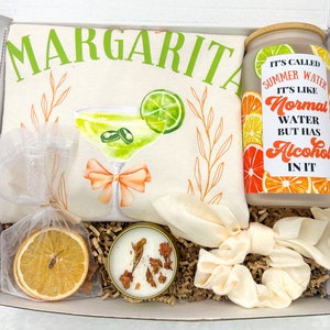 May include: A gift box filled with a white tote bag with a margarita illustration and the text "Margarita", a candle with dried orange slices, a clear glass jar with a citrus design and the text "It's called Summer Water. It's like Normal Water but has Alcohol in it", and a white scrunchie.