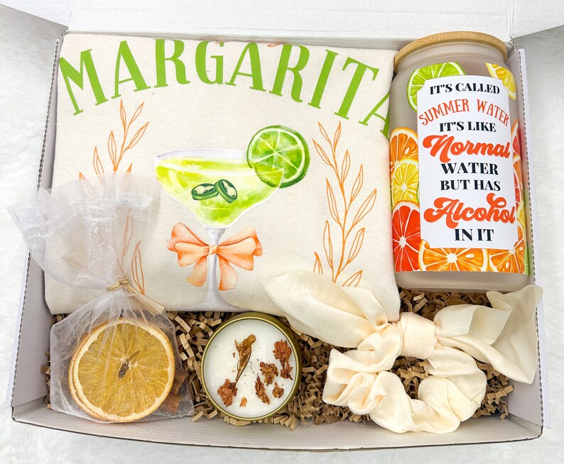 May include: A gift box filled with a white tote bag with a margarita illustration and the text "Margarita", a candle with dried orange slices, a clear glass jar with a citrus design and the text "It's called Summer Water. It's like Normal Water but has Alcohol in it", and a white scrunchie.