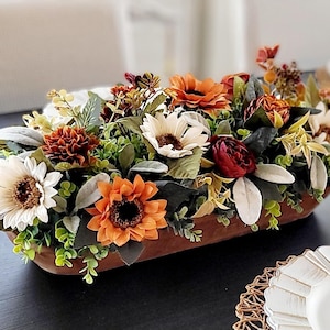 May include: A long, oval-shaped wooden dough bowl filled with an arrangement of artificial flowers in autumn colors, including cream, orange, and deep red sunflowers and small blossoms, accented with various green leaves and foliage sprigs.