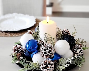 May include: A circular centerpiece featuring a central white pillar candle surrounded by blue and white spherical ornaments, frosted pinecones, and green evergreen sprigs arranged on a black base, creating a festive holiday display.