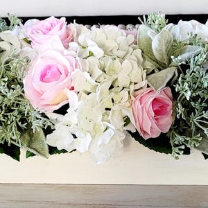 May include: A white wooden planter box filled with a floral arrangement of pink roses and white hydrangeas. The arrangement is accented with green foliage.