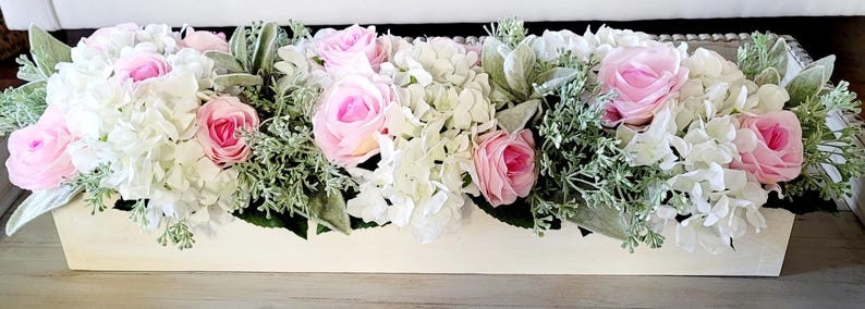 May include: A white wooden planter box filled with a floral arrangement of pink roses and white hydrangeas. The arrangement is accented with green foliage.