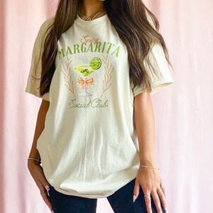 May include: A beige t-shirt with a graphic of a margarita glass with a lime wedge and the text "Spicy Margarita Social Club".