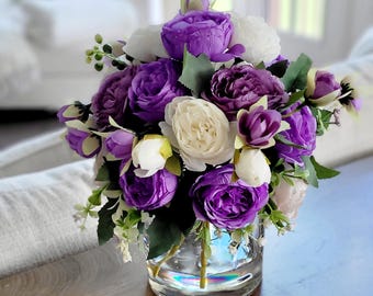 May include: A floral arrangement featuring artificial purple and white roses and small buds in a clear glass vase. The bouquet includes green leaves and stems, with the flowers densely packed to create a rounded, multi-layered display.