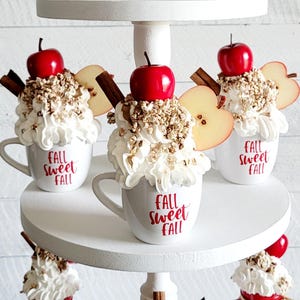 Può includere: Tre tazze in ceramica bianca con panna montata, bastoncini di cannella e fette di mela rossa. Le tazze sono decorate con il testo "Fall Sweet Fall" in lettere rosse.