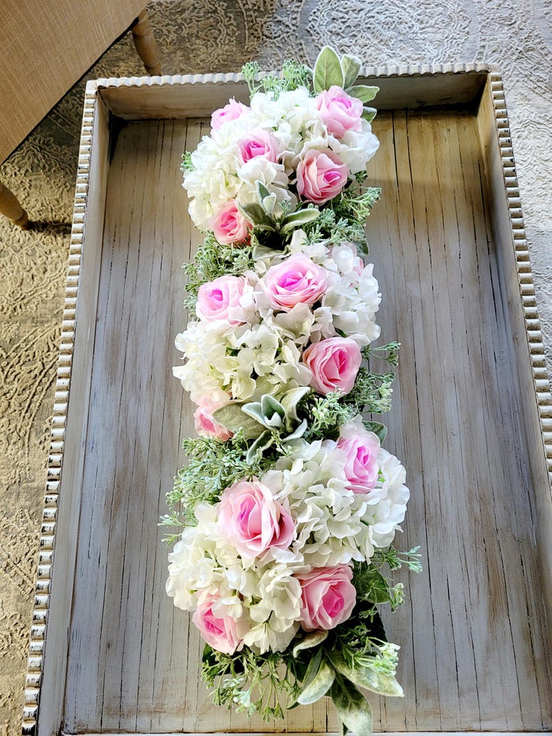 May include: A long, rectangular tray with a whitewashed wood finish and a beaded edge. The tray is filled with a floral arrangement of pink roses and white hydrangeas with green foliage.
