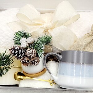 May include: A white gift box filled with a white mug, a small white planter with a pine sprig and pine cones, a white towel, a gold spoon, and a small burlap bag.
