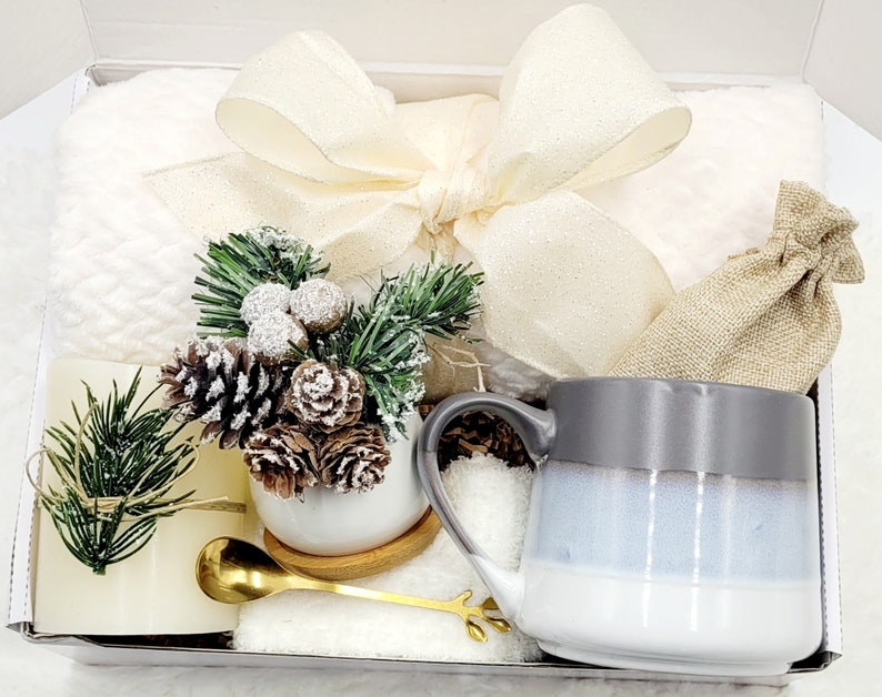 May include: A white gift box filled with a white mug, a small white planter with a pine sprig and pine cones, a white towel, a gold spoon, and a small burlap bag.