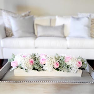 May include: A white wooden box filled with a floral arrangement of pink and white flowers. The flowers are arranged in a long, rectangular box and are sitting on a white coffee table.