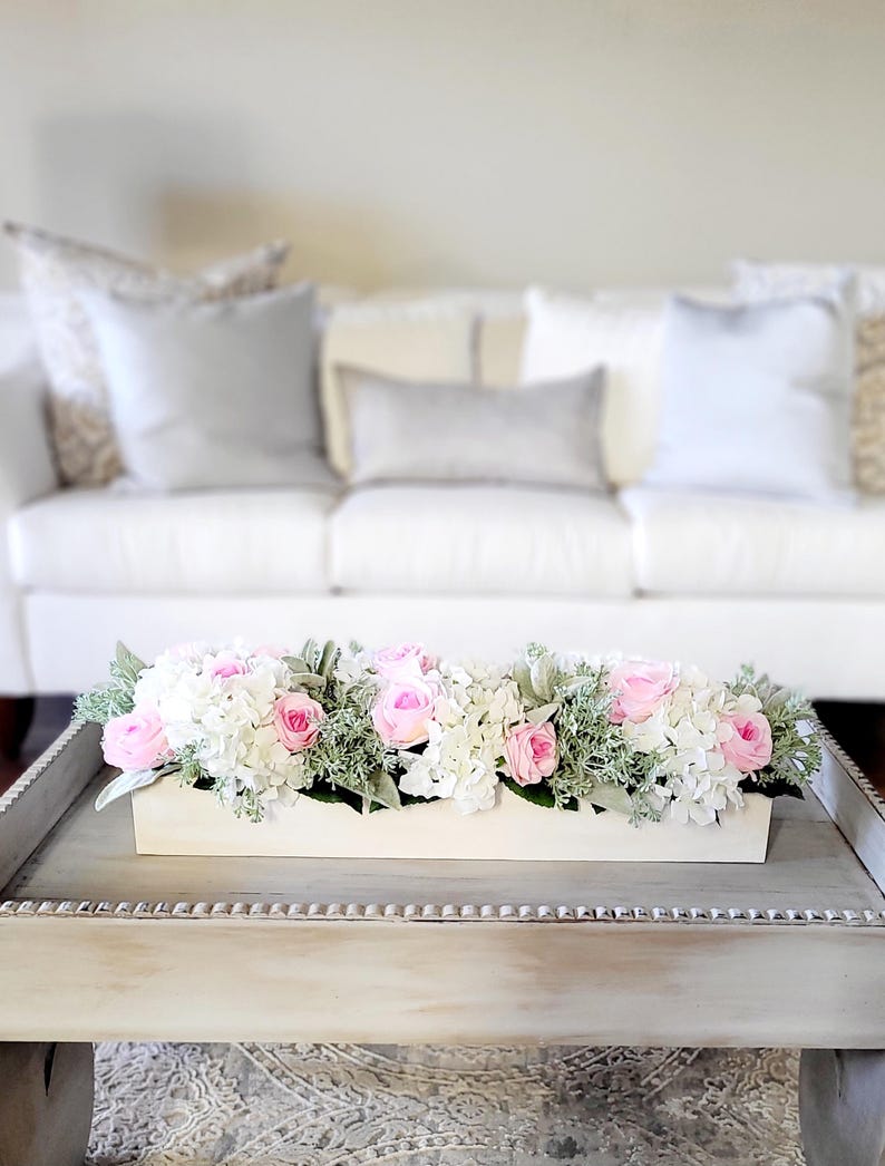 May include: A white wooden box filled with a floral arrangement of pink and white flowers. The flowers are arranged in a long, rectangular box and are sitting on a white coffee table.