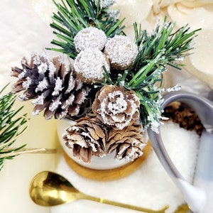 May include: A white ceramic mug with a brown wooden base, filled with a small pine tree, pine cones, and white berries. The mug is surrounded by a white fluffy blanket and a white satin ribbon.