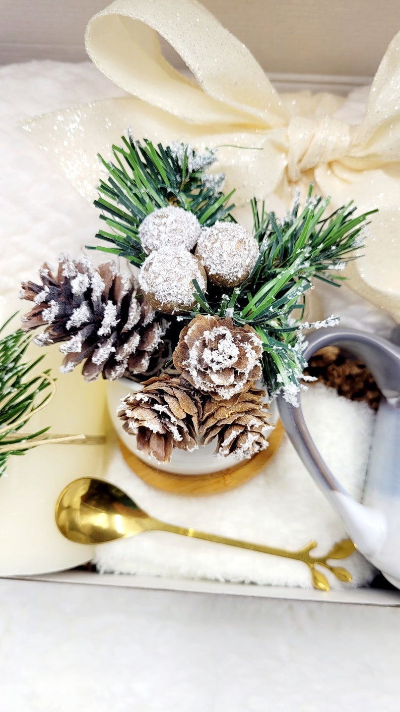 May include: A white ceramic mug with a brown wooden base, filled with a small pine tree, pine cones, and white berries. The mug is surrounded by a white fluffy blanket and a white satin ribbon.