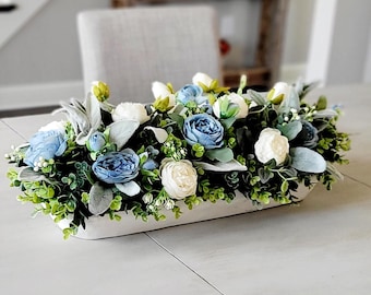 May include: A long, rectangular white wooden dough bowl filled with an arrangement of artificial flowers and greenery. The display features light blue and cream-colored roses, small white buds, and fuzzy, sage-green lamb's ear leaves.