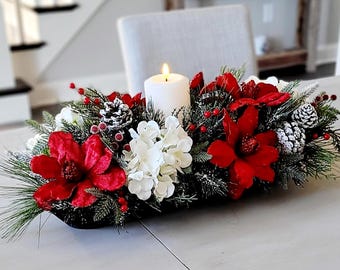 May include: A holiday centerpiece featuring a white pillar candle nestled in a black dough bowl filled with red magnolia flowers, white hydrangea blossoms, frosted pinecones, green pine needles, and small red berries.