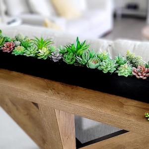 May include: A long, black rectangular planter box filled with a variety of artificial succulents in shades of green, pink, and purple. The planter sits on a wooden console table. Trailing green plants cascade down the sides. A gold lamp is visible on the left.