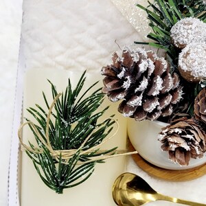 May include: A white candle with a green pine sprig tied with twine, a small white ceramic pot with a pine cone and a gold spoon on a white surface.