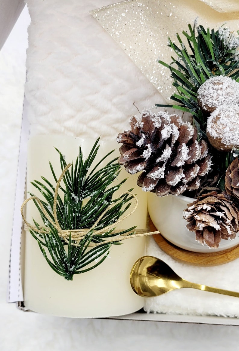 May include: A white candle with a green pine sprig tied with twine, a small white ceramic pot with a pine cone and a gold spoon on a white surface.