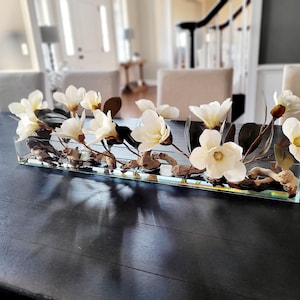 May include: A rectangular glass vase filled with water and driftwood, holding a bouquet of white magnolia flowers.