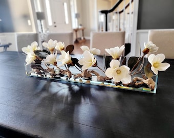 Puede incluir: Un jarrón de vidrio rectangular lleno de agua y madera flotante, que contiene un ramo de flores de magnolia blancas.