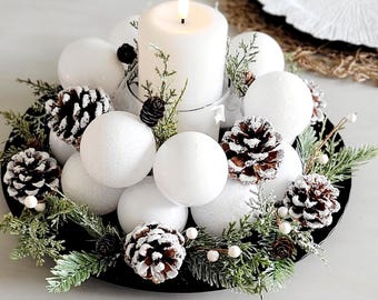 May include: A holiday centerpiece featuring a central white pillar candle in a glass holder, surrounded by white spherical ornaments, brown pinecones with white-tipped edges, and green evergreen sprigs with small white berries on a black tray.