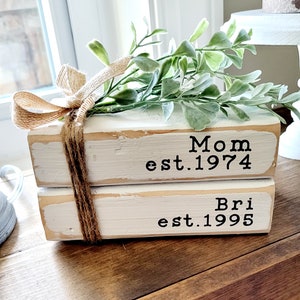 May include: Two white painted wooden blocks stacked on top of each other. The top block has the text "Mom est. 1974" and the bottom block has the text "Bri est. 1995". The blocks are decorated with a brown twine bow and a sprig of green leaves.