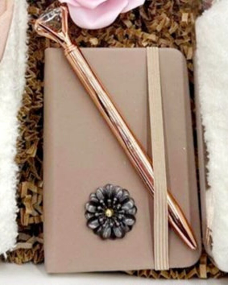 May include: A small brown leather journal with a rose gold pen and a decorative flower embellishment.
