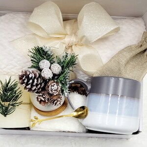 May include: A white gift box filled with a white fluffy blanket, a gray and white mug, a small burlap bag, a small potted evergreen tree with pine cones, a gold spoon, and a white candle.