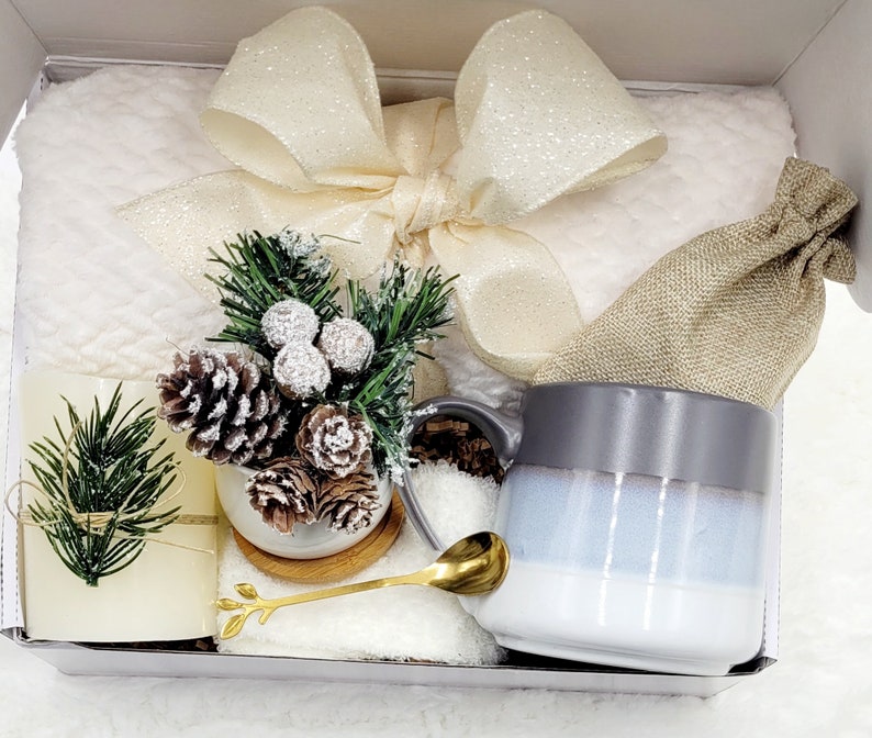 May include: A white gift box filled with a white fluffy blanket, a gray and white mug, a small burlap bag, a small potted evergreen tree with pine cones, a gold spoon, and a white candle.