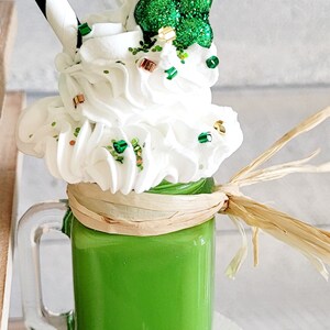 May include: A green milkshake in a glass jar with a white lid and a black and white striped straw. The milkshake is topped with whipped cream and green glitter, and a green shamrock.