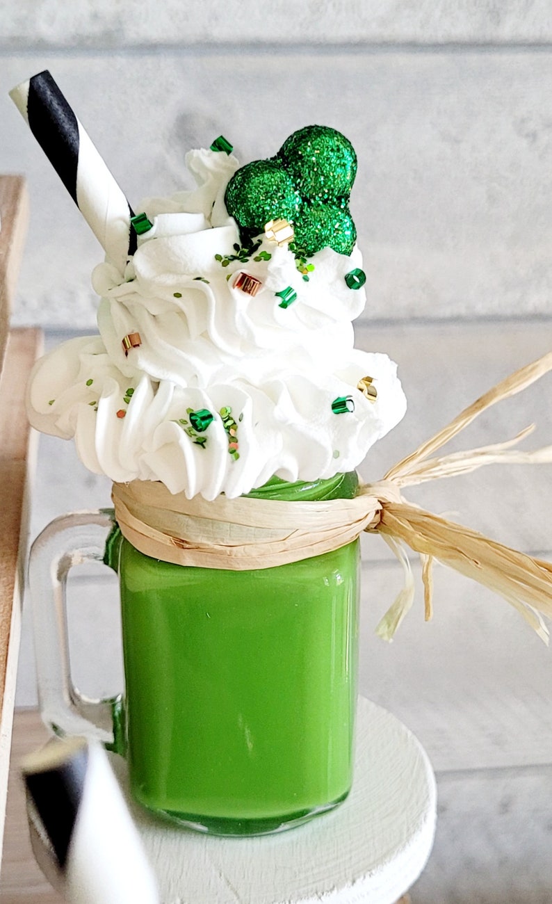 May include: A green milkshake in a glass jar with a white lid and a black and white striped straw. The milkshake is topped with whipped cream and green glitter, and a green shamrock.