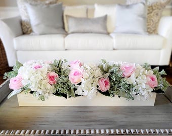 May include: A white rectangular planter filled with a floral arrangement of pink roses and white hydrangeas. The planter is sitting on a grey wooden table.