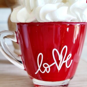 May include: Red glass mug with a white handle and the word "love" written in white with a heart shape. The mug is topped with white whipped cream.