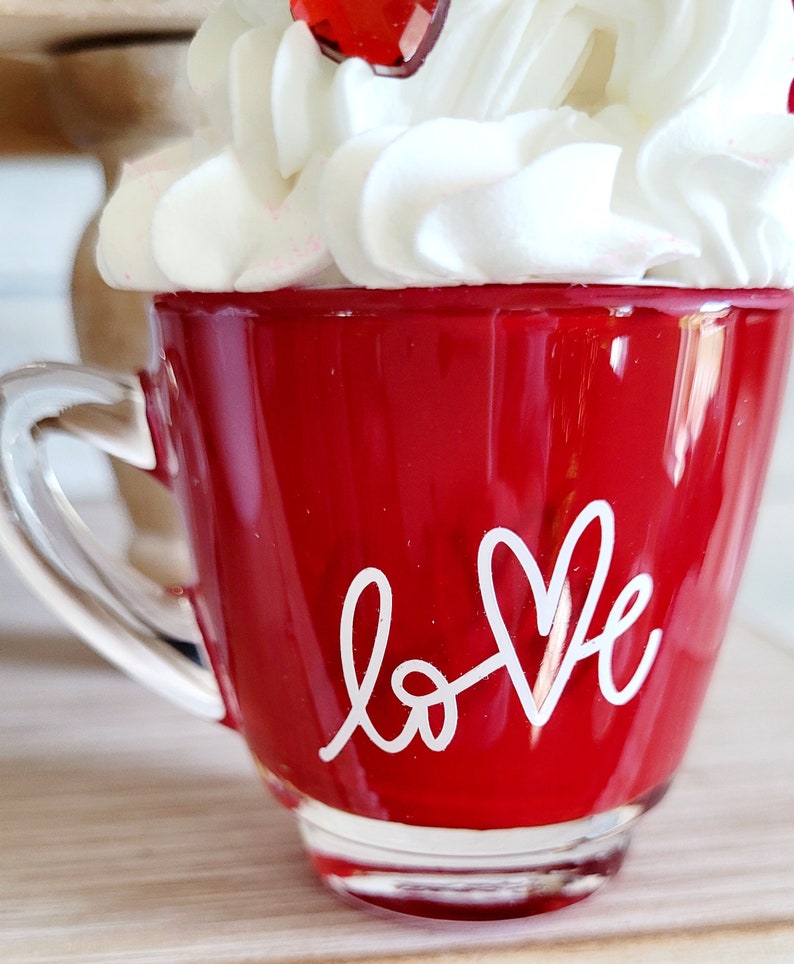 May include: Red glass mug with a white handle and the word "love" written in white with a heart shape. The mug is topped with white whipped cream.