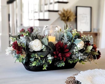 May include: A black oval bowl filled with artificial flowers including white roses and deep red dahlias, accented with green eucalyptus leaves and small sprigs. A clear glass candle holder sits in the center containing a small lit pillar candle.